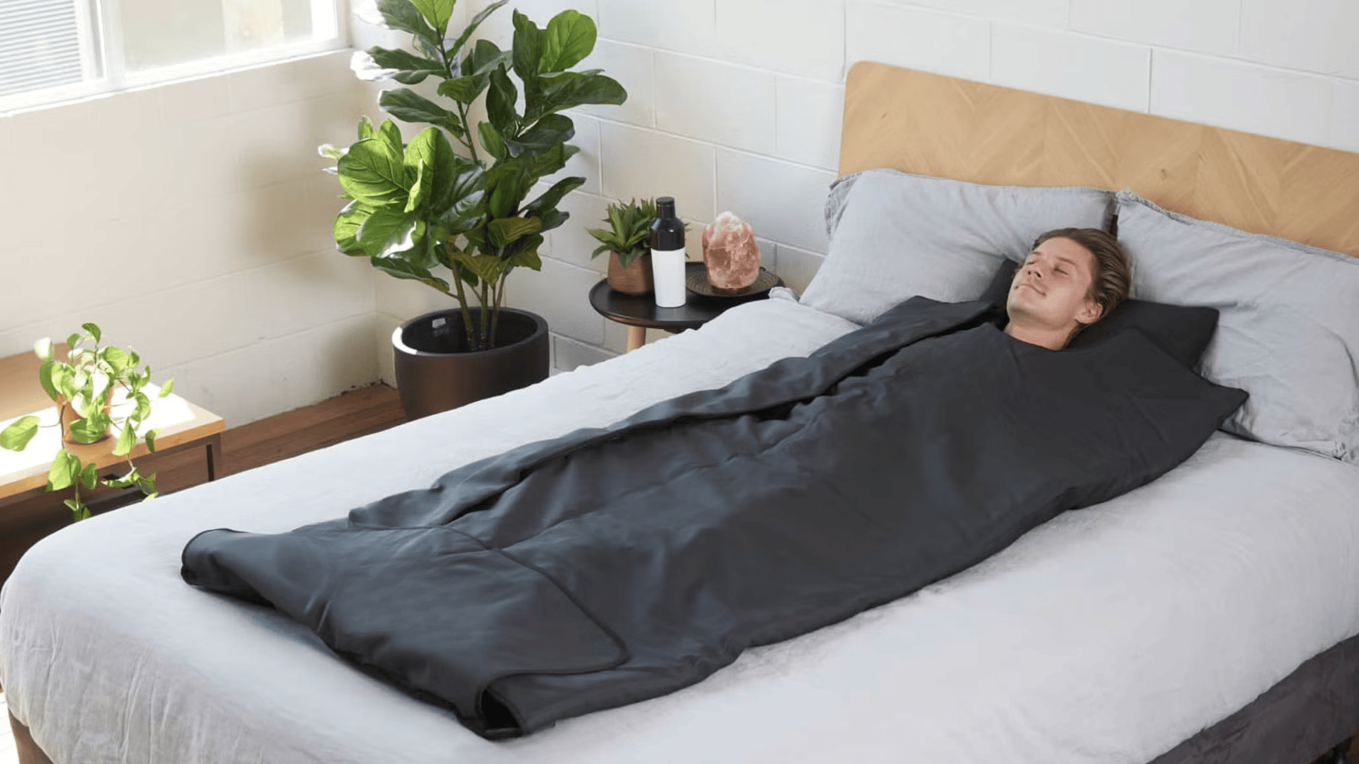 Person relaxing inside a black infrared sauna blanket on a bed in a bright, minimal bedroom with plants