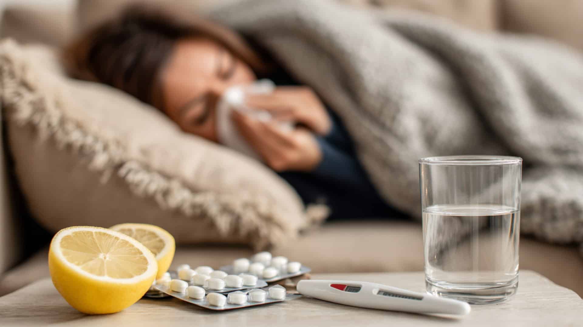 Sick woman resting under a blanket with tissues, lemon, tablets, thermometer, and water on a table in the foreground, indoors.