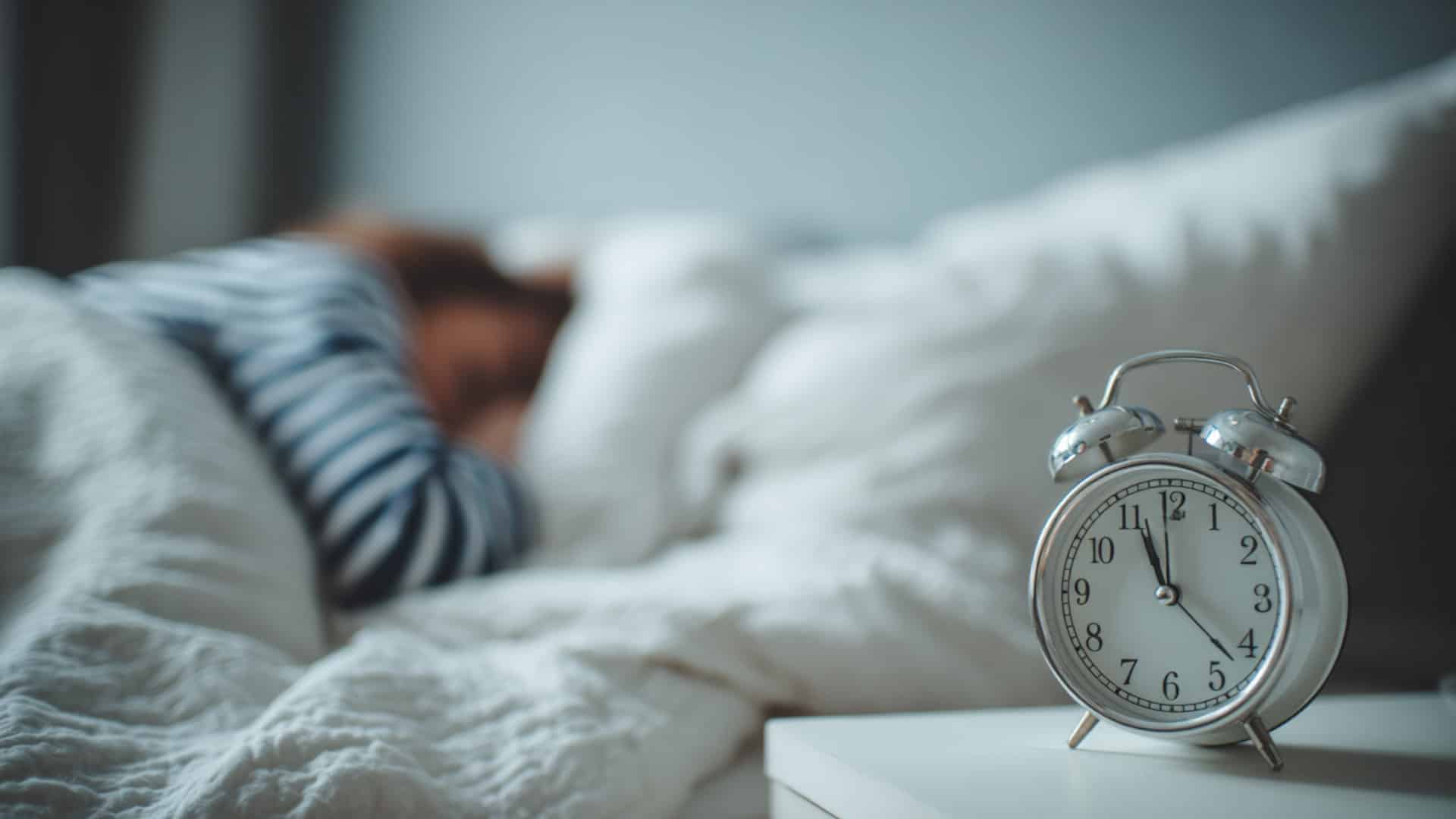 White alarm clock on bedside table in focus, with a person sleeping under white blankets blurred softly in the background of the home