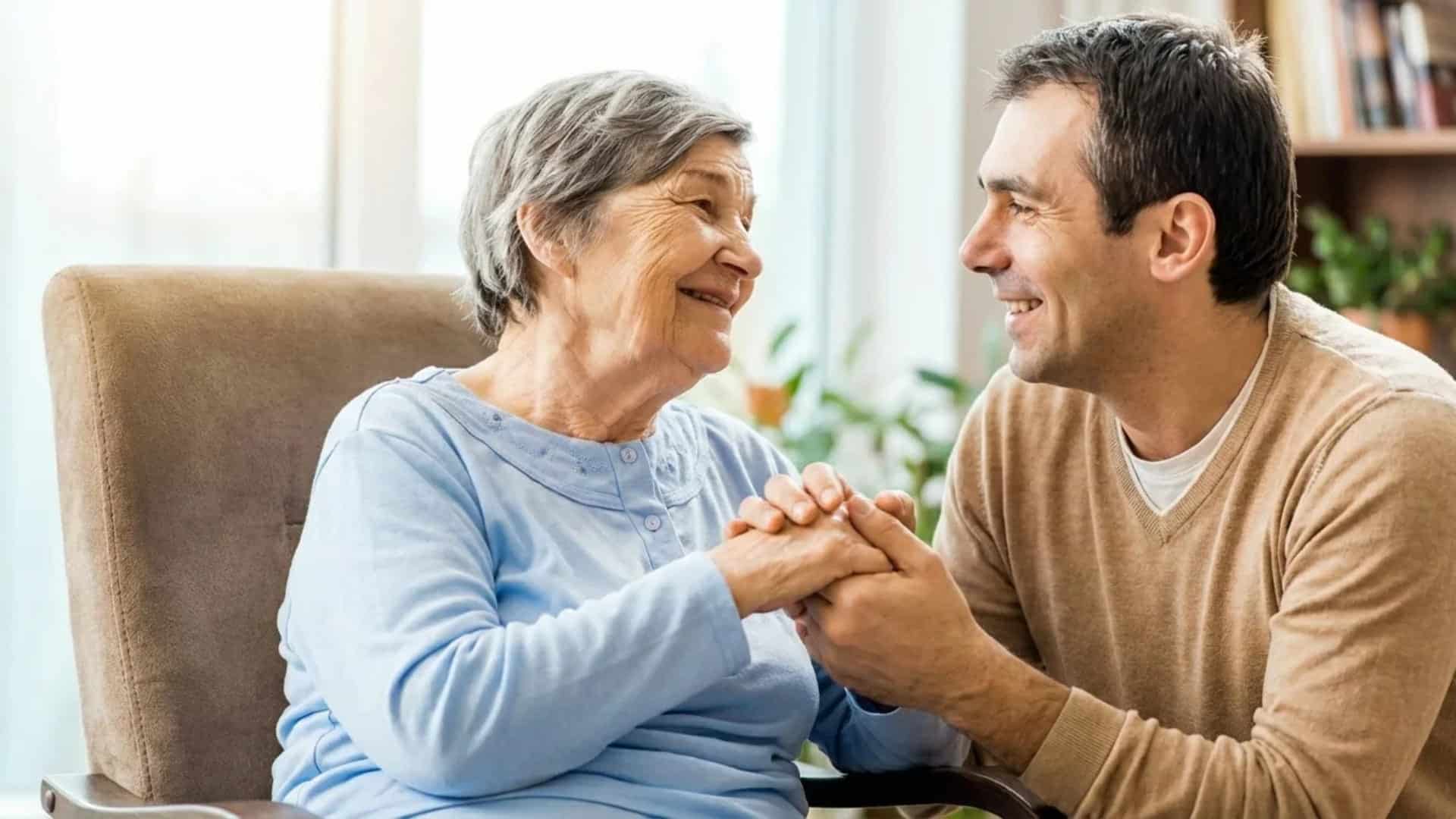 Elderly woman smiling and holding hands with a male caregiver in a bright room, showing warmth, trust, comfort, and support