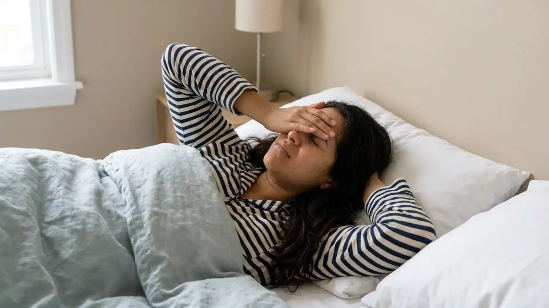 Woman lying in bed with hand on forehead, showing tired or uneasy feeling during sleep.