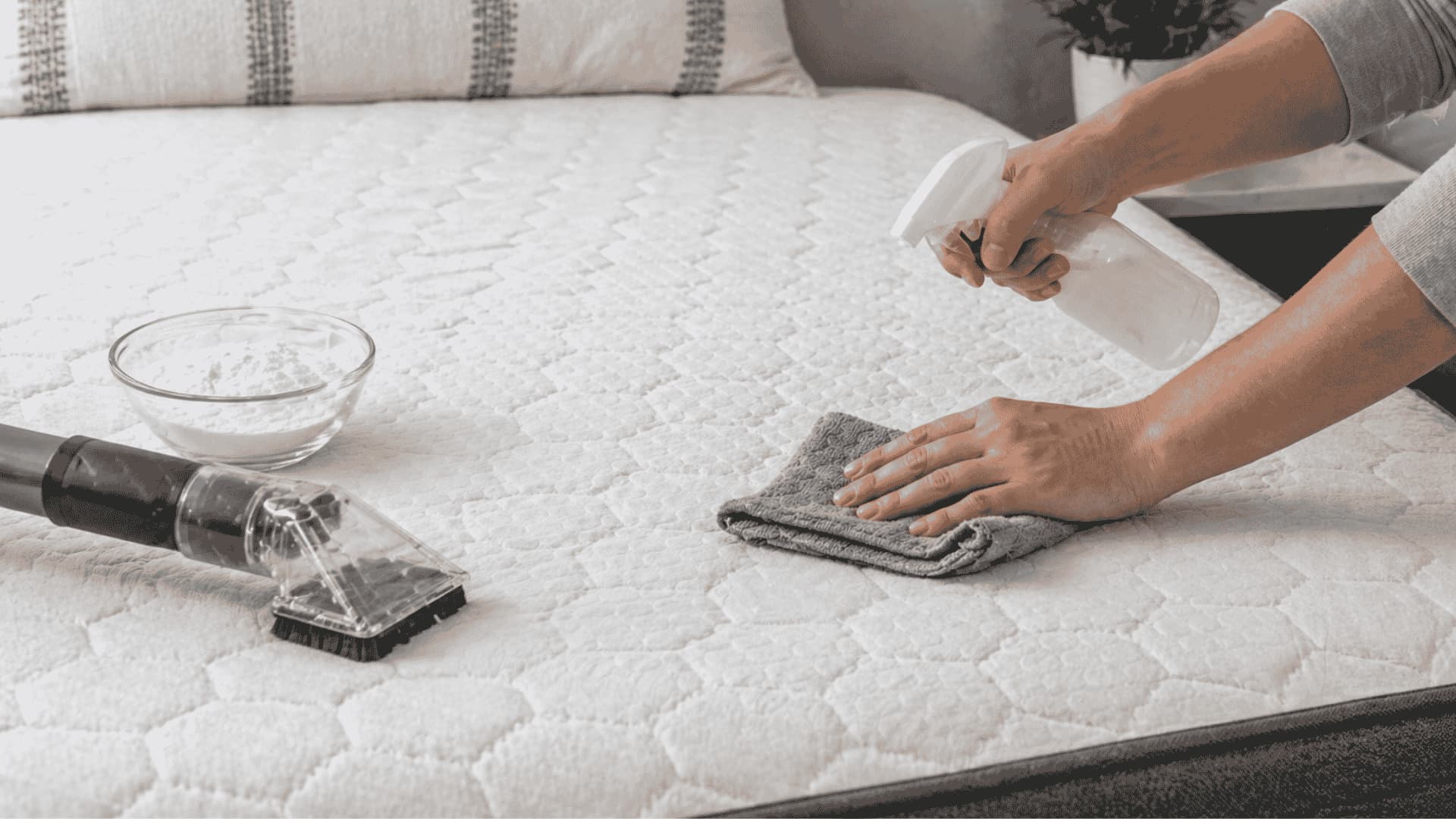 Person cleaning a memory foam mattress using spray bottle, cloth, and vacuum in a bright bedroom setting