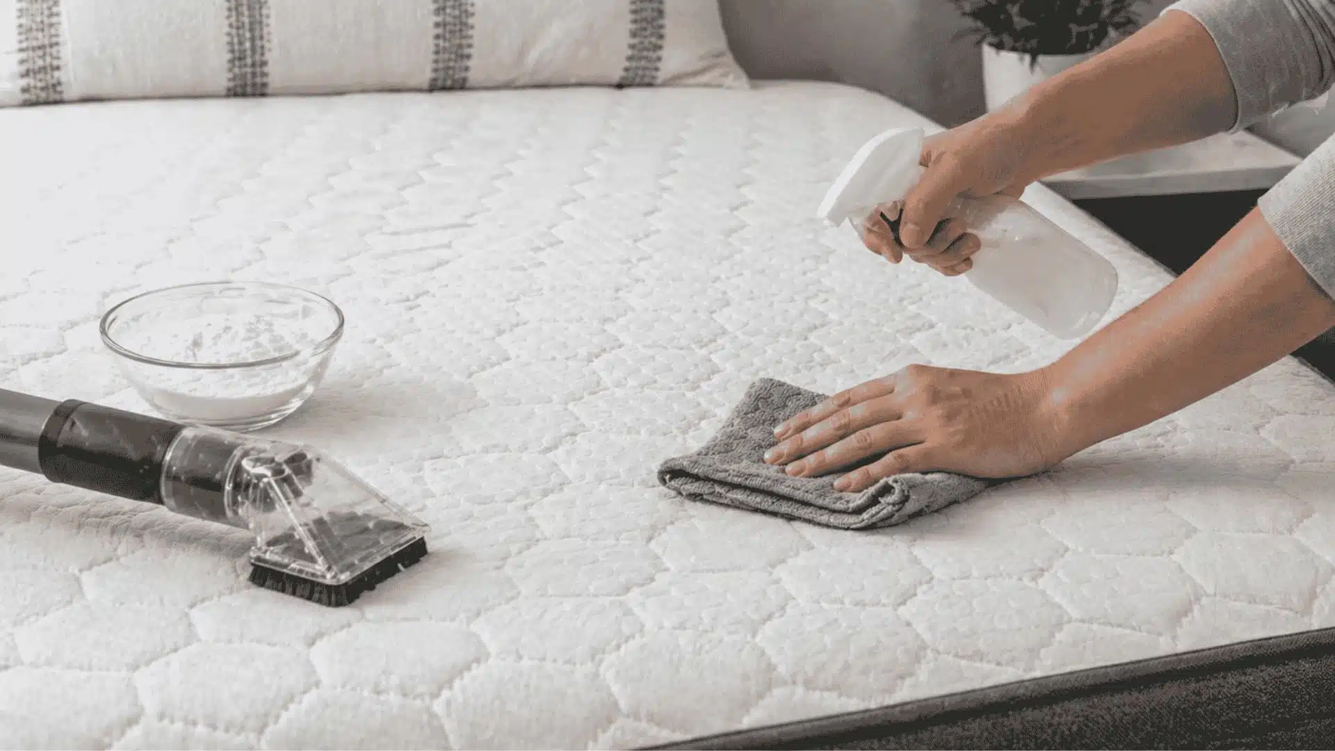 Person cleaning a memory foam mattress using spray bottle, cloth, and vacuum in a bright bedroom setting