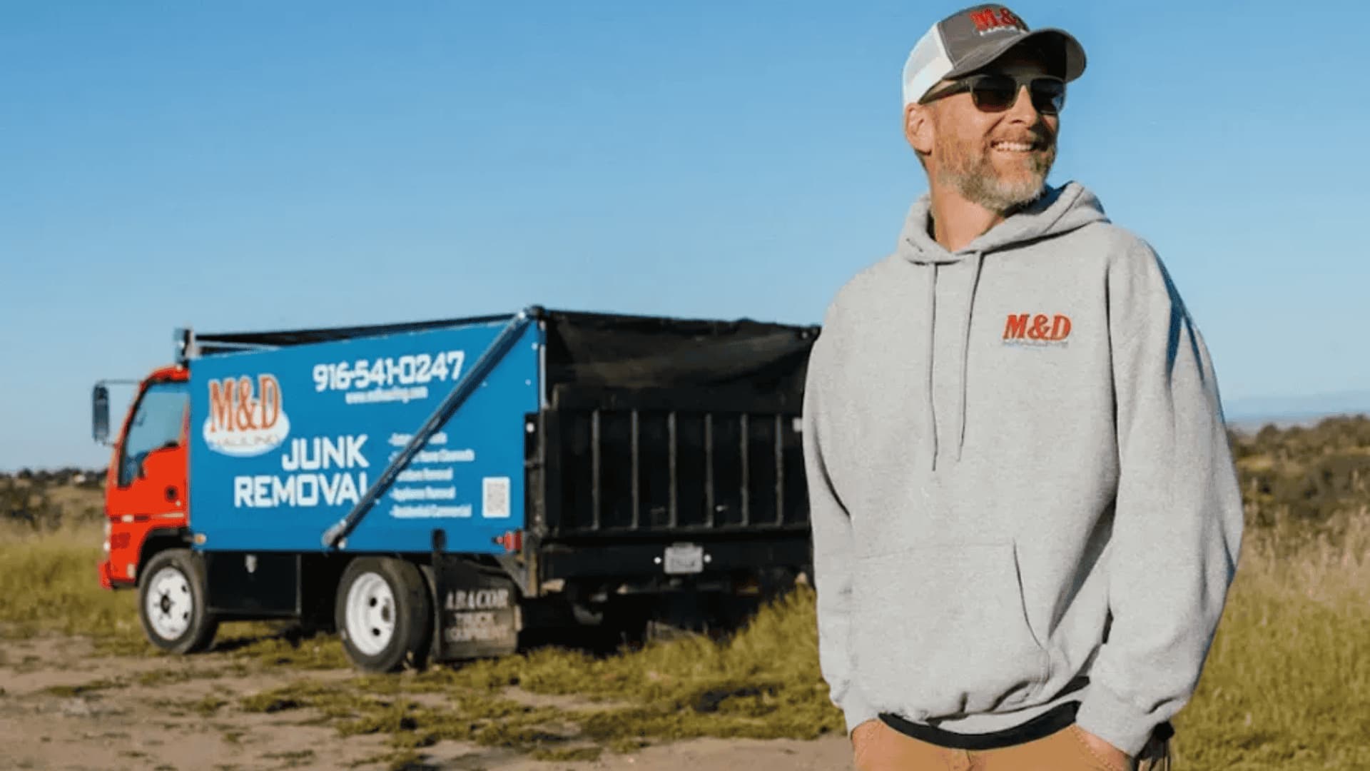 Man in M&D Hauling hoodie and cap standing in front of a junk removal truck in an open field.