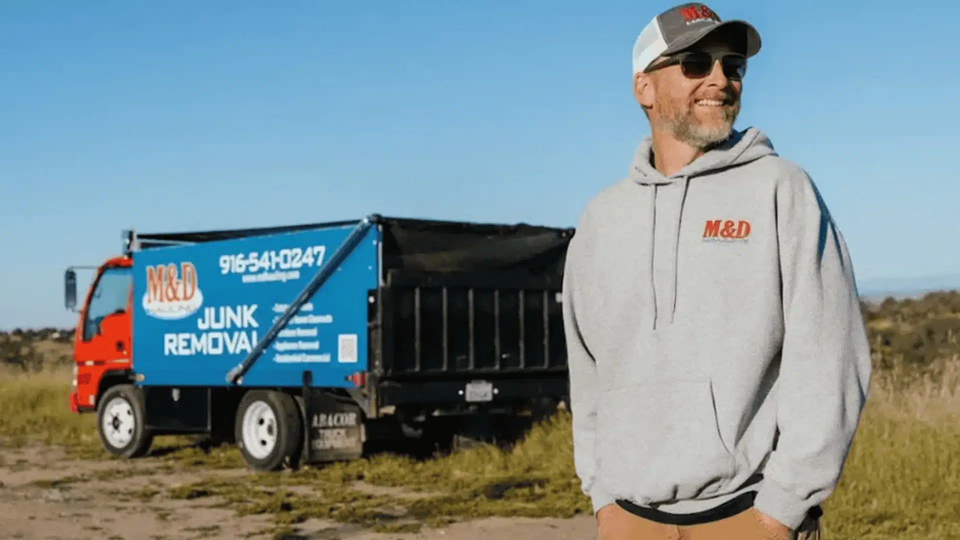 Man in M&D Hauling hoodie and cap standing in front of a junk removal truck in an open field.