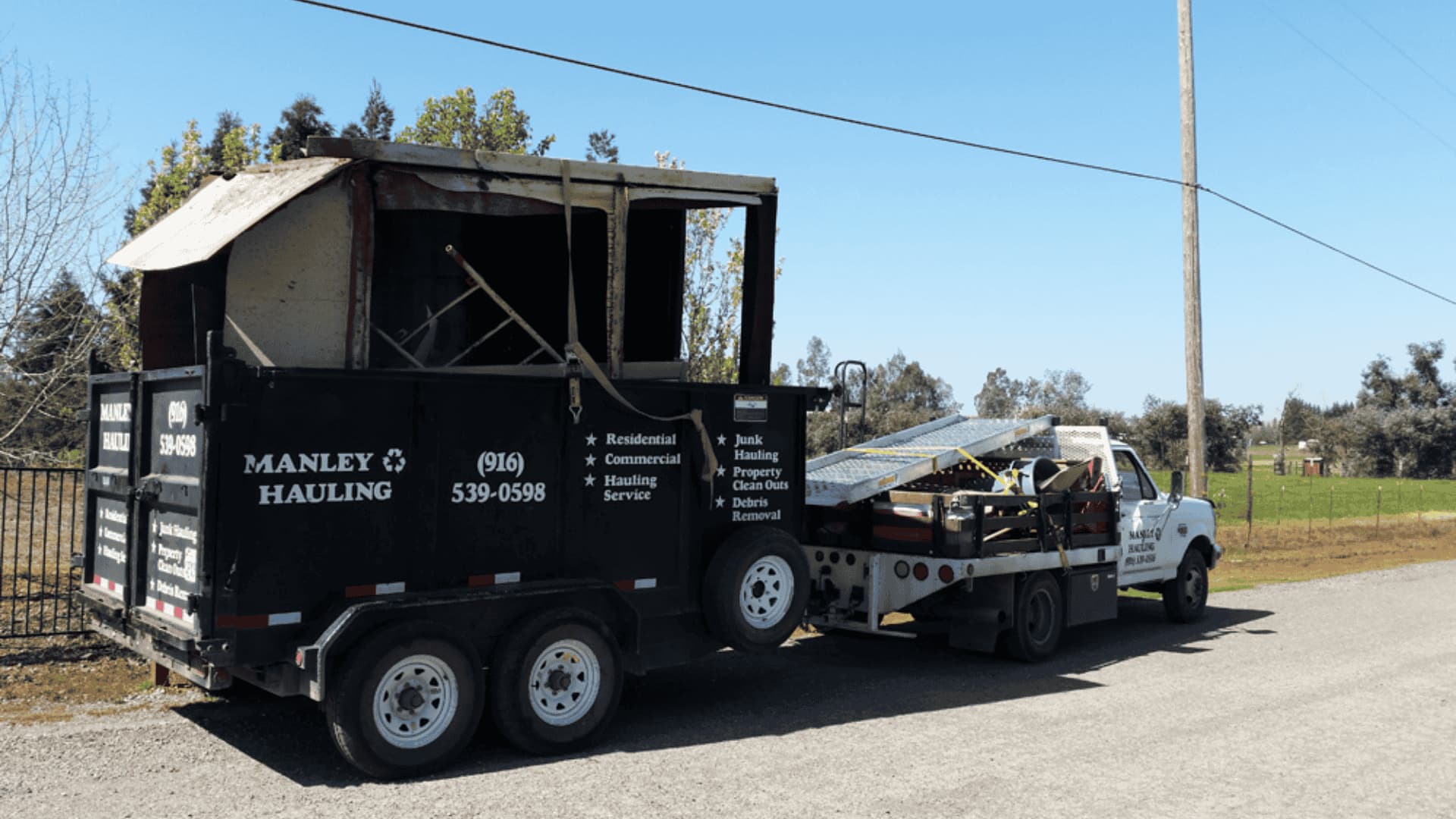 Manley Hauling dump trailer loaded with debris attached to a pickup truck on a rural road.
