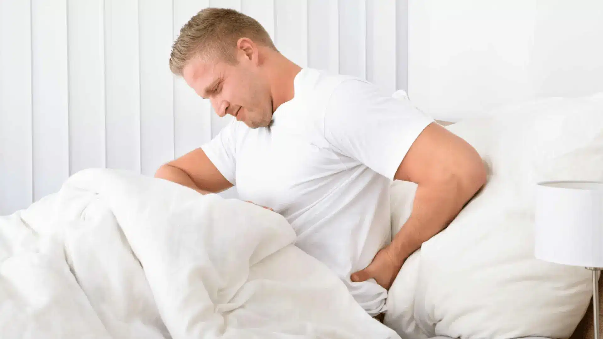 a man sitting up in bed, grimacing and holding his lower back in pain, surrounded by white pillows and linens
