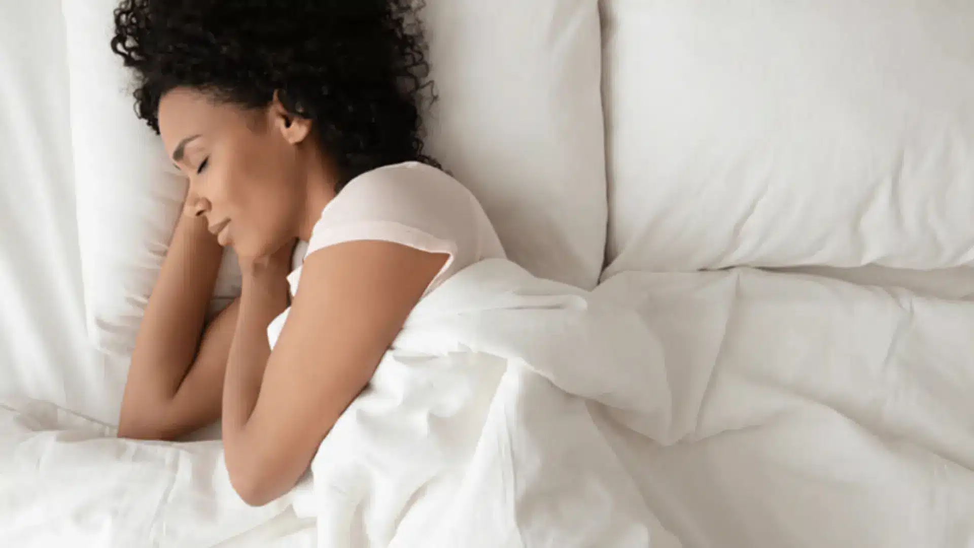 a woman with curly hair sleeping peacefully on her side in a bed with bright, clean white linens