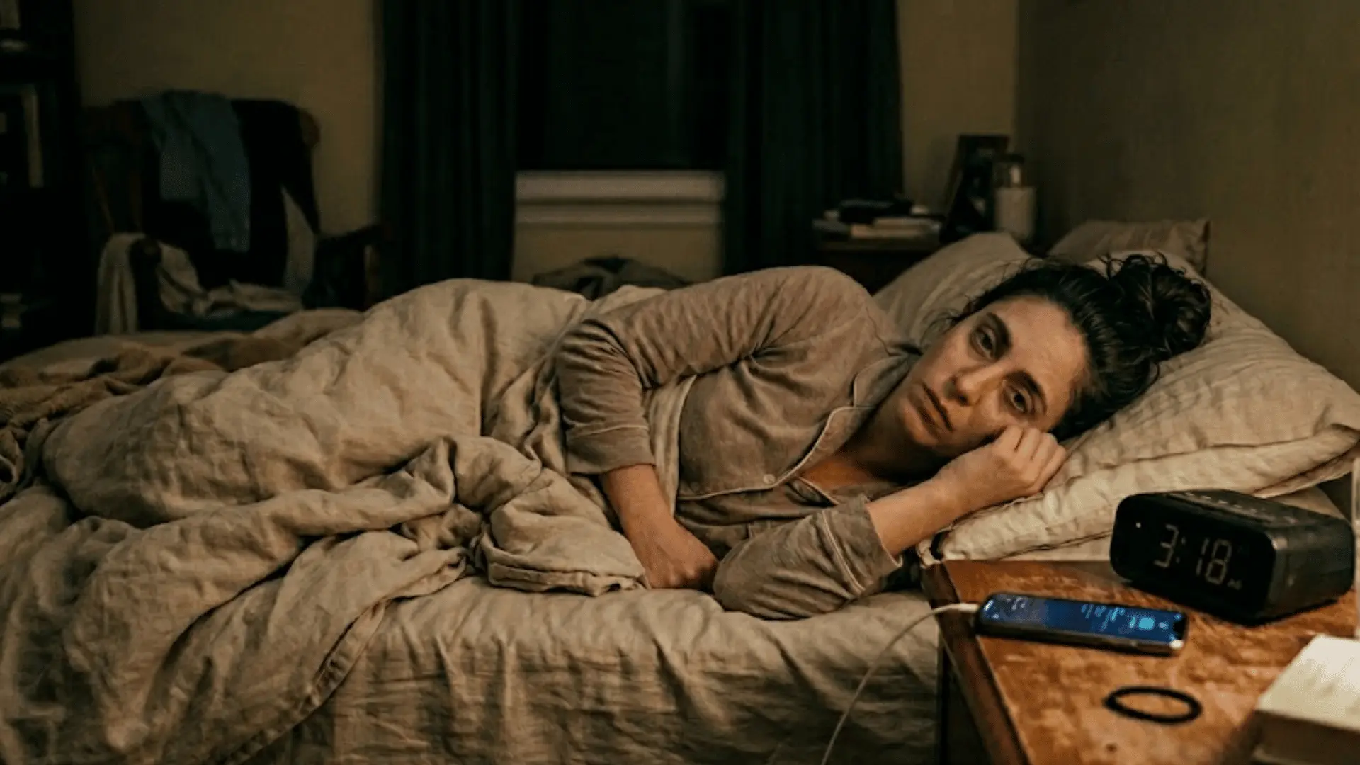 A wide-awake woman lies in bed looking tired next to a digital alarm clock showing 3:18 AM in a dark room.