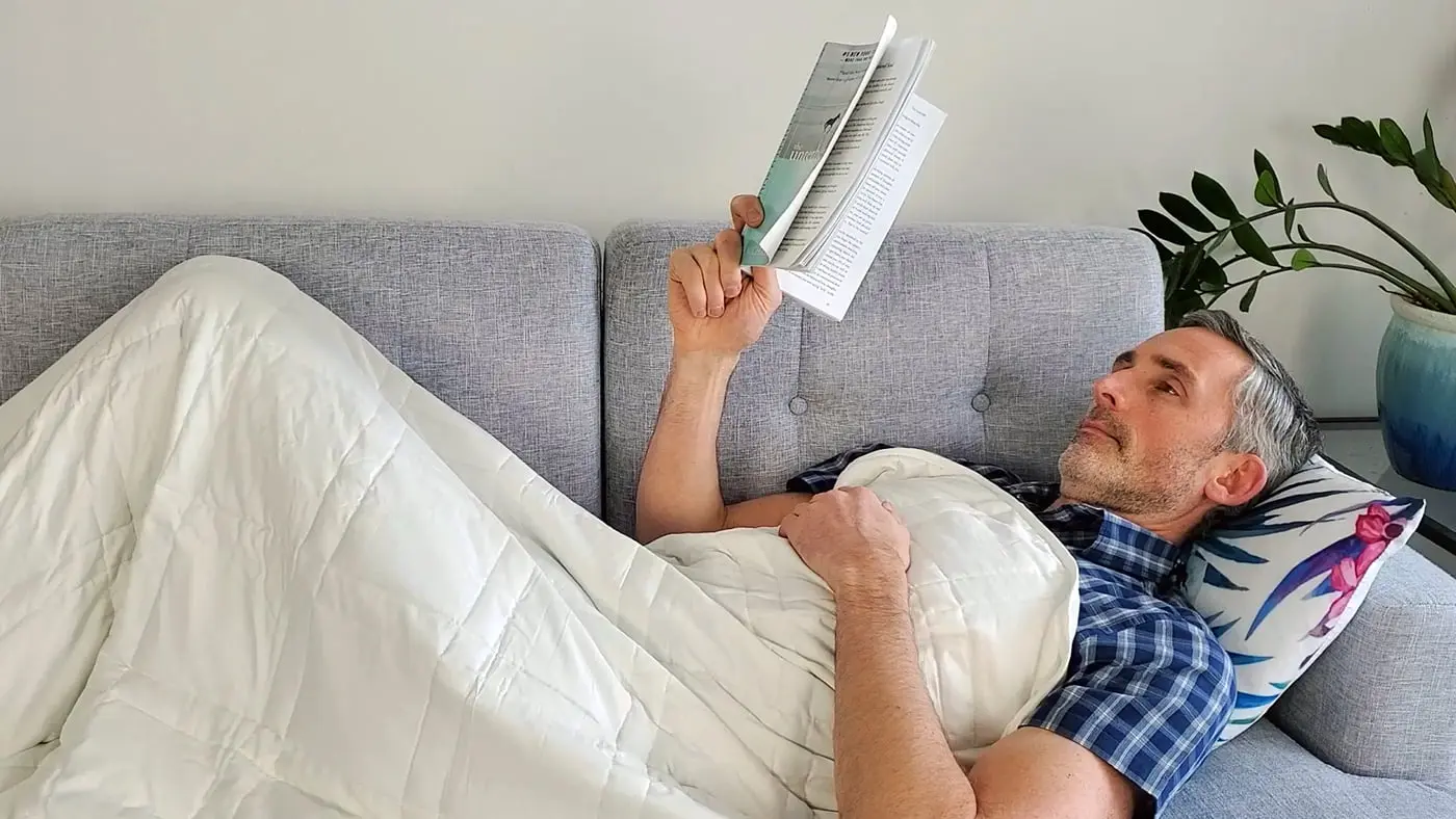Older man lying in bed under white weighted blanket reading a book