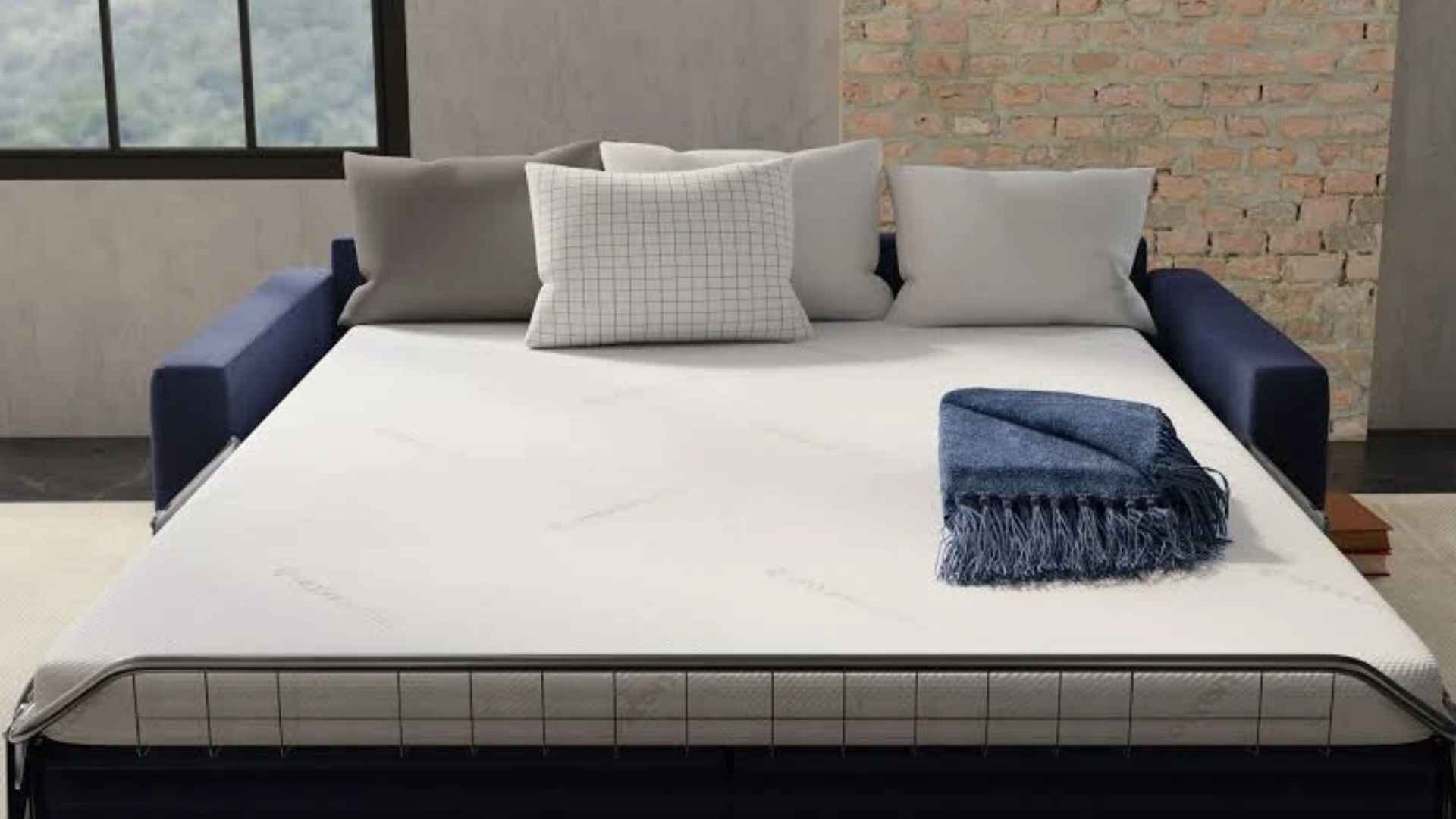 Modern industrial-style living room with a sofa bed, white mattress, pillows, and a folded blue blanket on top, brick wall and window in background.