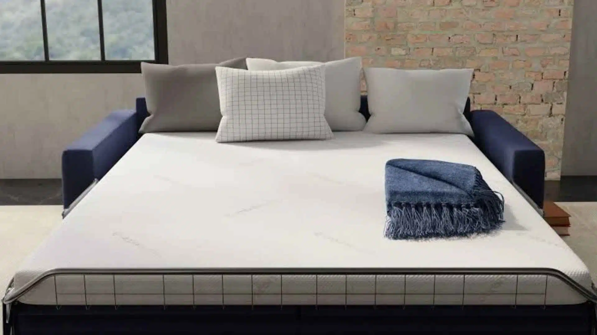 Modern industrial-style living room with a sofa bed, white mattress, pillows, and a folded blue blanket on top, brick wall and window in background.
