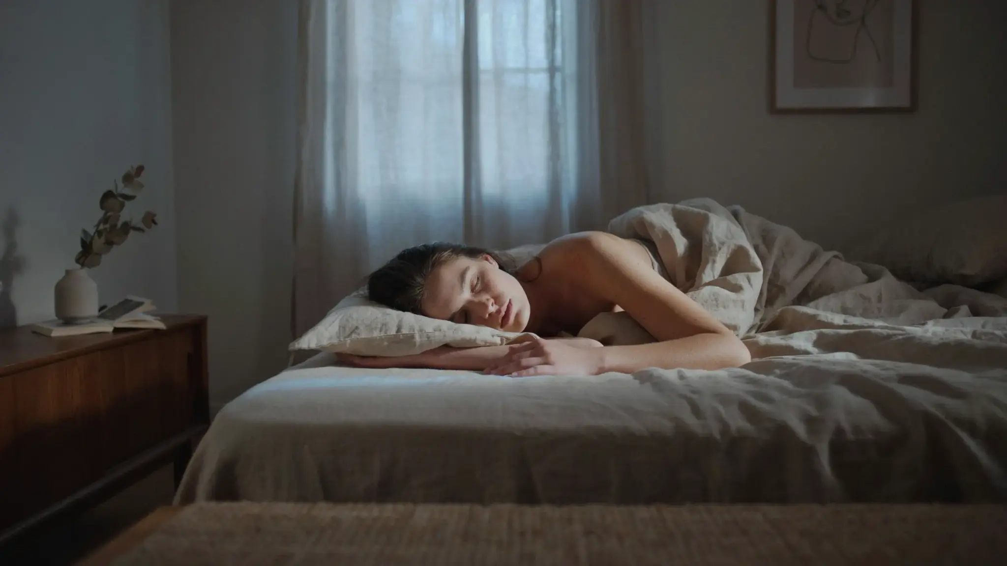 Person sleeping peacefully in a dimly lit bedroom at night, resting under soft bedding with moonlight filtering through sheer curtains.