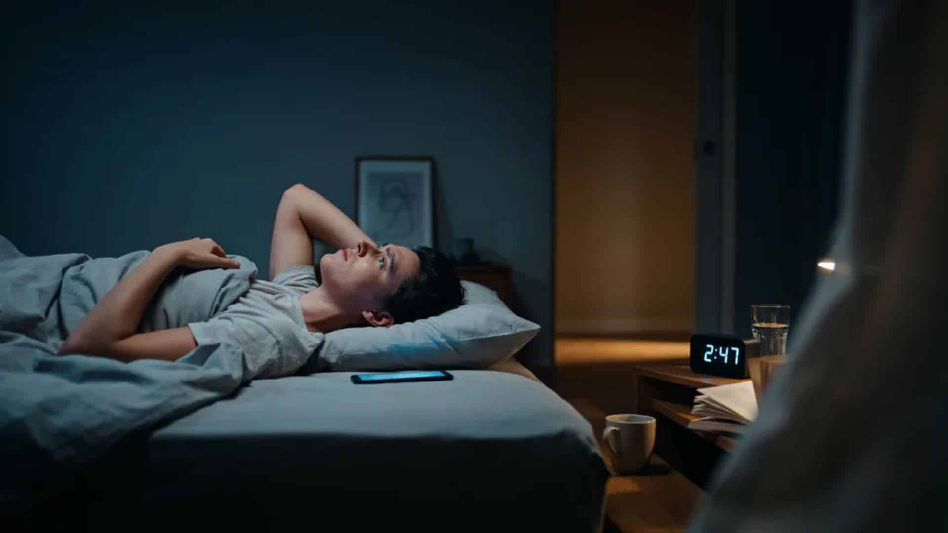 person lying awake in bed at night looking frustrated with phone nearby and clock showing late hour
