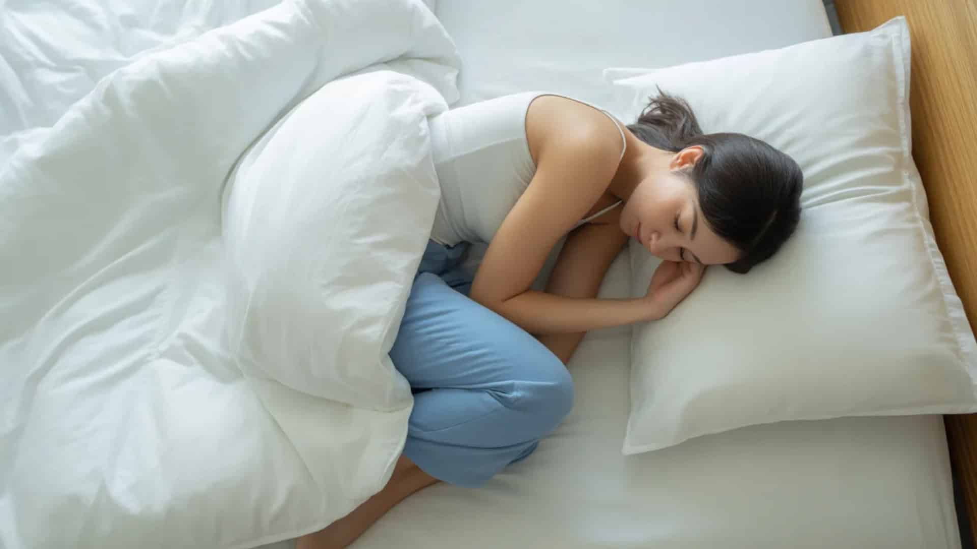 Woman asleep on her side in white bedding with bent knees and pillow nearby rest