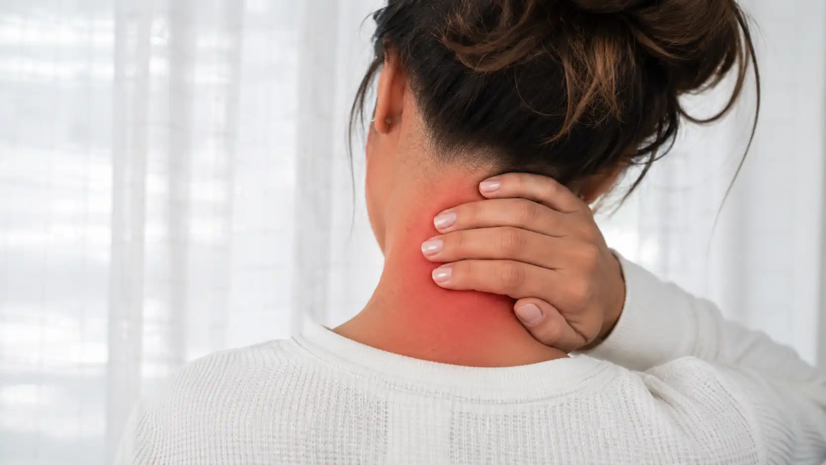 Rear view of a woman holding her neck, showing redness and discomfort associated with a pinched nerve, in a softly lit indoor setting.
