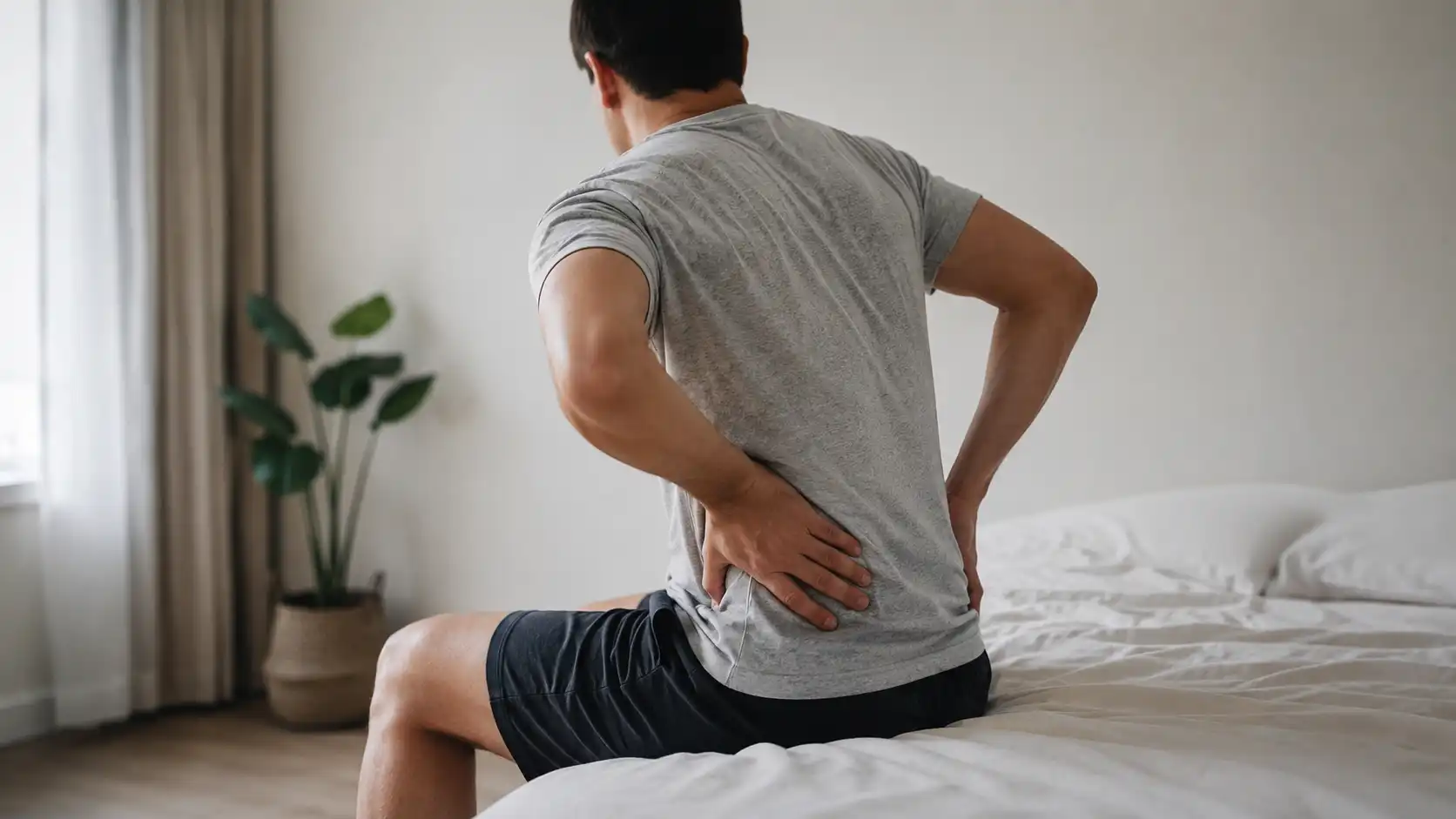 Man sitting on bed holding lower back in pain, showing discomfort in a modern bedroom setting.