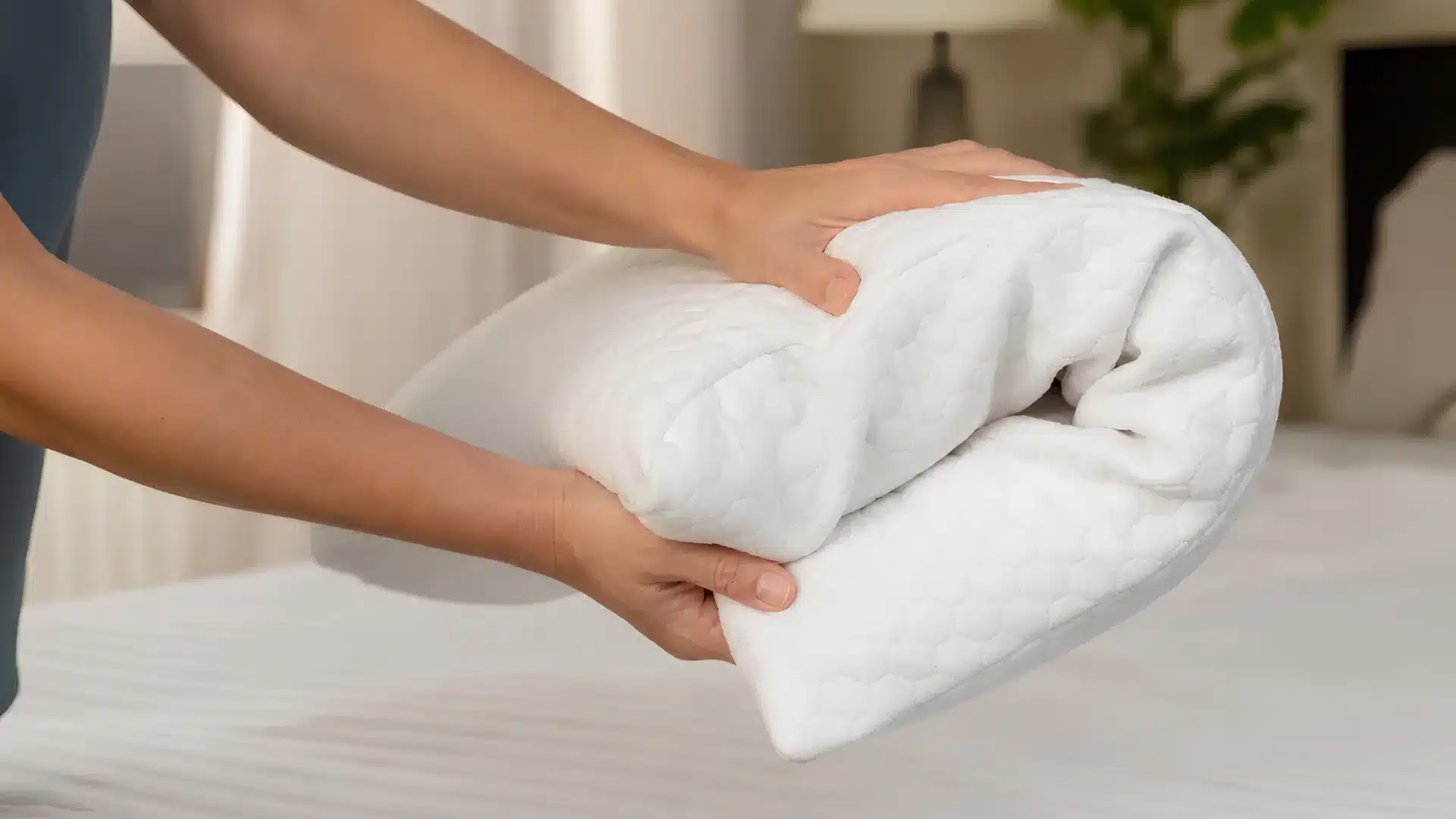 Hands folding a soft, quilted white mattress topper on a bed.