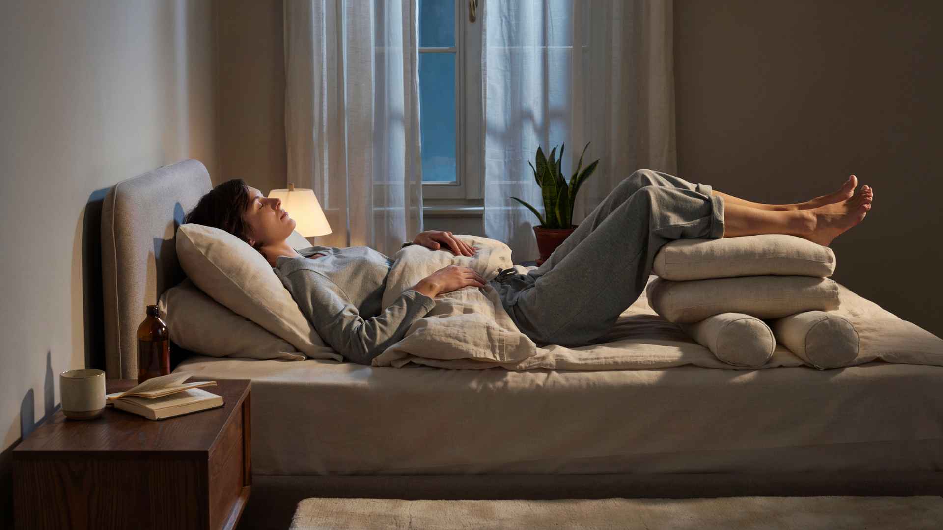 Person lying on bed with legs elevated on stacked pillows in a softly lit bedroom, resting comfortably at night.