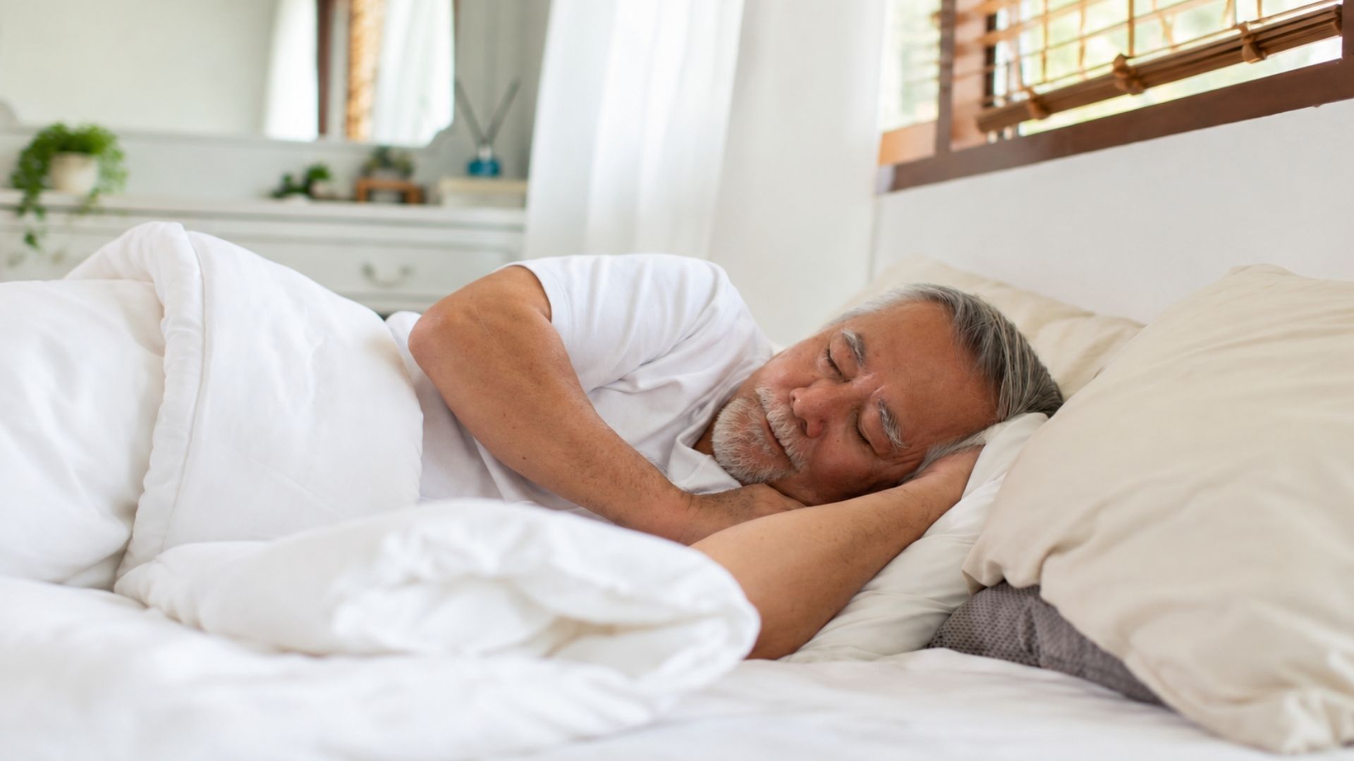 An older man sleeping comfortably in a bright bedroom with white bedding, showing a peaceful sleep environment for finding the ideal sleeping temperature.
