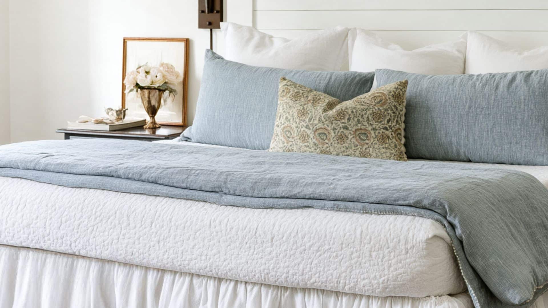 A neatly made bed styled with white bedding, dusty blue pillows and throw, and a patterned accent pillow