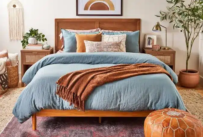 Bedroom with a wooden bed, blue and rust bedding, cane nightstands with lamps, and decor including a macrame hanging and potted plants