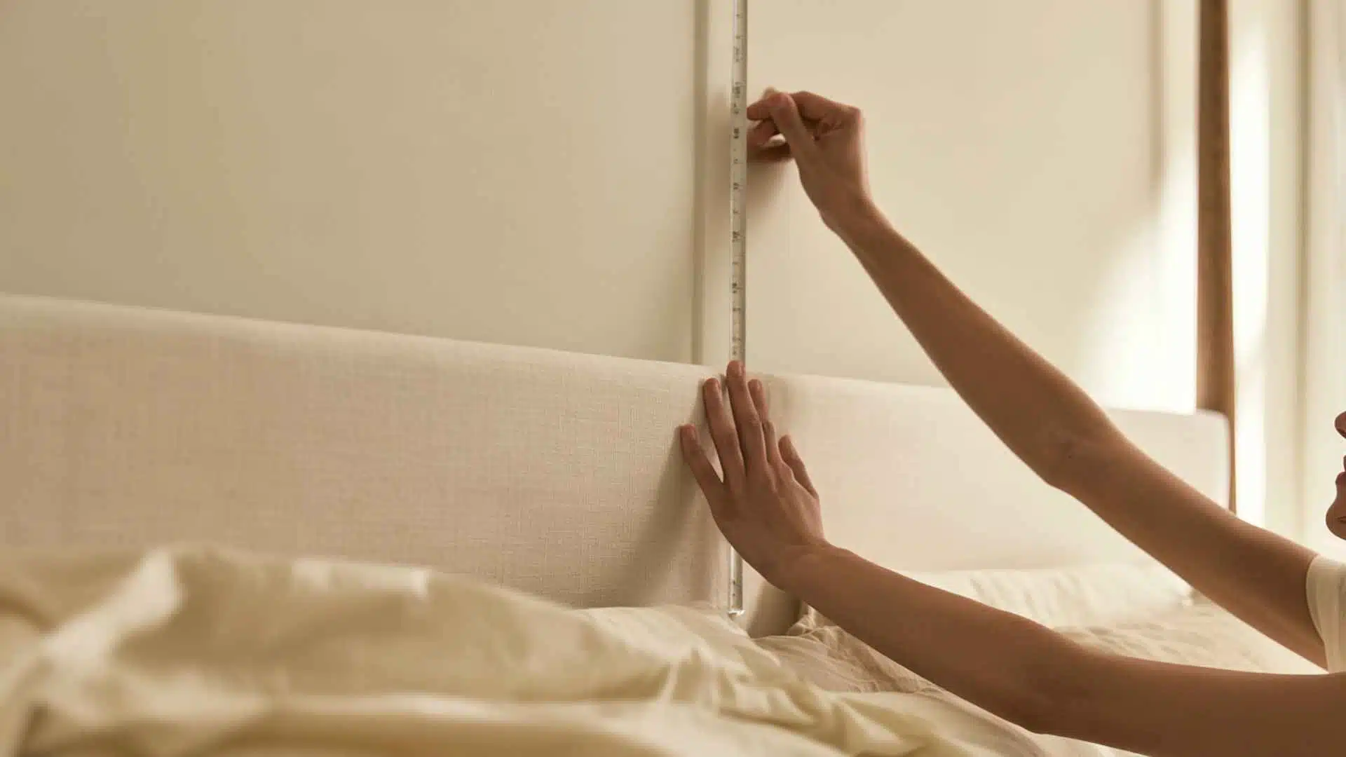 Woman measuring the wall and bed area with a tape measure before installing a DIY bed canopy