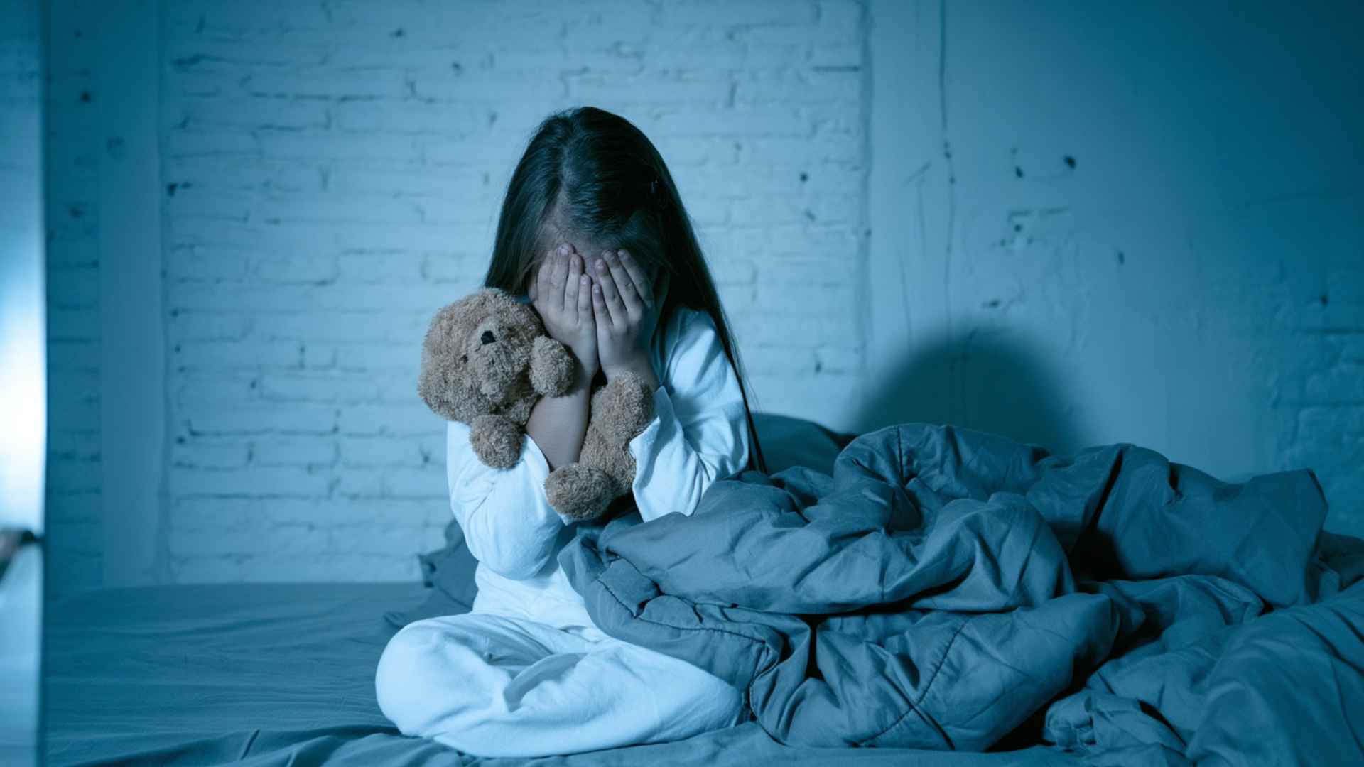 Young girl sitting awake in bed at night, covering her face while holding a teddy bear, showing fear of sleep.